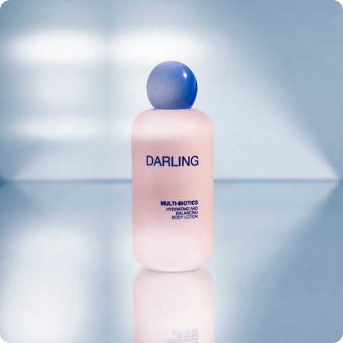 Roze flacon van Darling Multi-Biotics Body Lotion met een opvallende blauwe bolvormige dop, staand op een spiegelende lichtblauwe ondergrond.