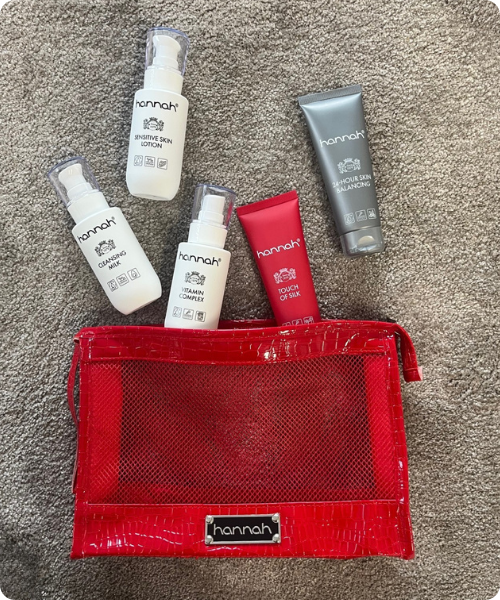 Een vijftal hannah huidverzorgingsproducten, waaronder cleansing milk, vitamin complex en touch of silk, liggend bij een rode doorschijnende toilettas op een grijze ondergrond.