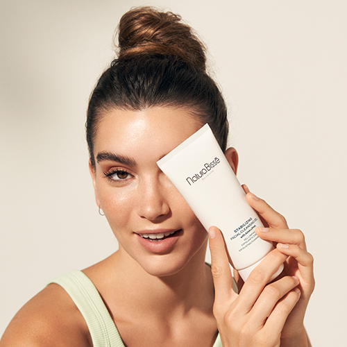 Een vrouw houdt de Natura Bissé Stabilizing Facial Cleansing Gel tube voor haar gezicht terwijl ze lacht.