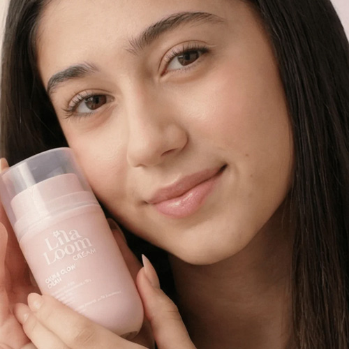 Een meisje houdt de lichtroze Lila Loom Calm and Glow Cream flacon tegen haar wang terwijl ze vriendelijk in de camera kijkt.