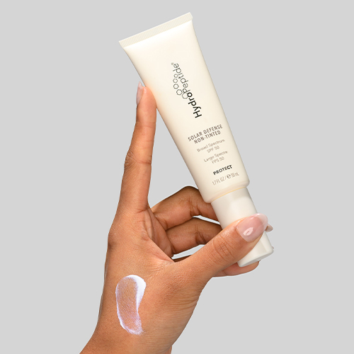 Hand houdt tube HydroPeptide Solar Defense Non Tinted SPF 50 vast met een beetje zonnebrandcrème op de huid