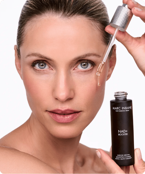 Close up van een model dat een glazen pipet met serum vasthoudt naast haar gezicht met daarnaast de fles van Marc Inbane NAD+ Booster.