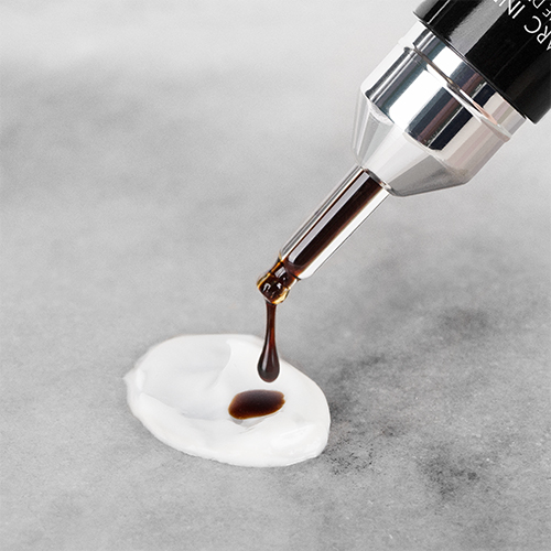 Een detailopname van een glazen applicator waaruit een donkerbruine zelfbruiner druppel valt op een veeg witte verzorgingscrème