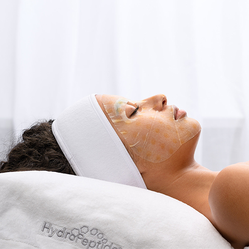 Een liggende persoon met het HydroPeptide Firming Vitamin C Mask op het gezicht tijdens een behandeling.