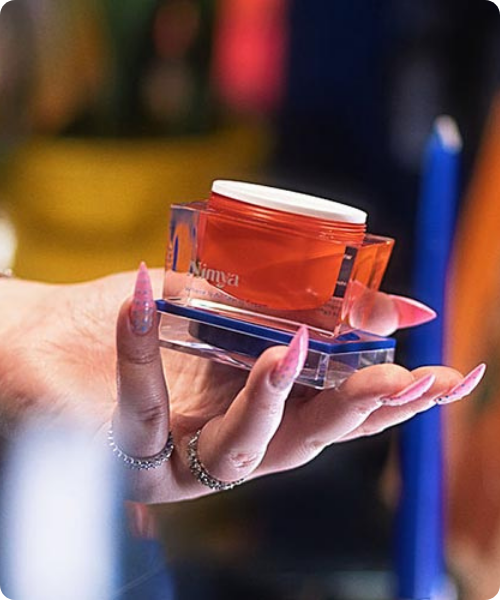Een hand met lange roze nagels houdt het oranje potje van Nimya Where It All Starts Cream vast op een transparante glazen houder.