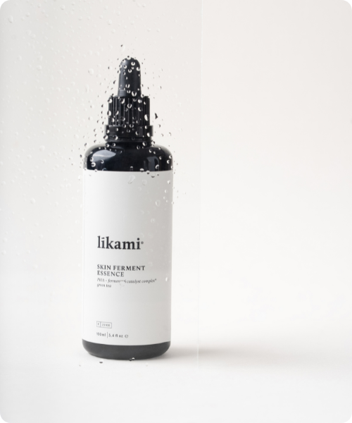 Zwarte glazen fles Likami Skin Ferment Essence met witte etiket en pipet achter een glas met waterdruppels