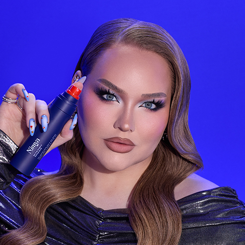 NikkieTutorials poseert met een blauwe flacon Nimya setting spray in haar hand tegen een blauwe achtergrond.