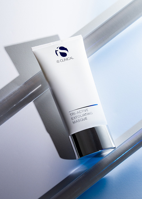 Een witte tube iS Clinical Tri-Active Exfoliating Masque, diagonaal geplaatst op een abstracte achtergrond met lichtblauwe en grijze accenten.