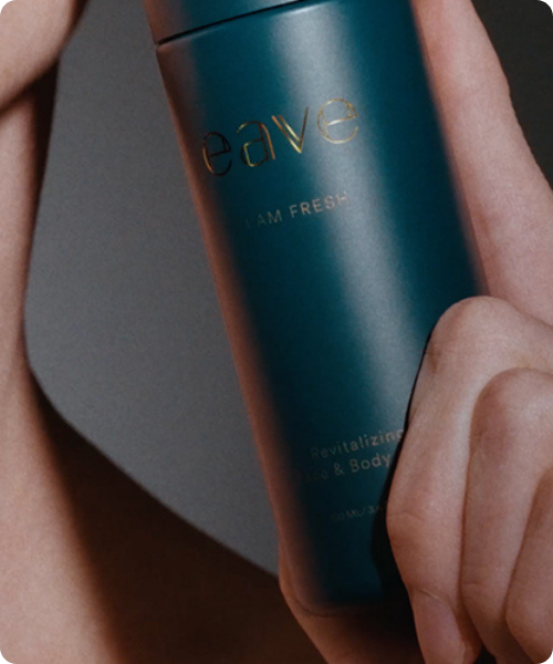 Close-up van een hand die een donkergroene flacon van EAVE 'I AM FRESH' Revitalizing Face & Body vasthoudt. De tekst op de flacon is in goud gedrukt.