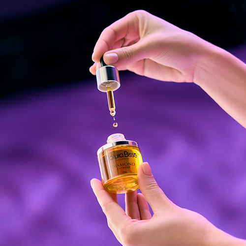 Handen die de pipet van de Diamond Extreme Oil boven het glazen flesje houden terwijl een druppel goudgele olie valt, tegen een paarse achtergrond.