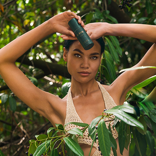 Een vrouw die poseert in een groene, bosrijke omgeving terwijl ze het EAVE I AM BRIGHT serum boven haar hoofd vasthoudt.