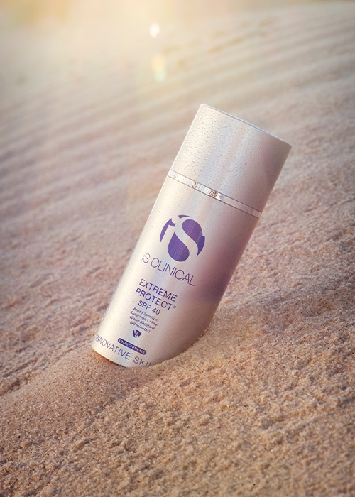 Een flesje iS CLINICAL Extreme Protect SPF 40 zonnebrandcrème, gedeeltelijk in het zand op een strand geplaatst met zonlicht op de achtergrond.
