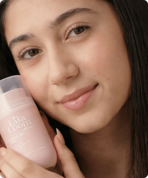 Een close-up van een glimlachende jonge vrouw die een roze flacon van Lila Loom 'Calm & Glow' crème tegen haar wang houdt.