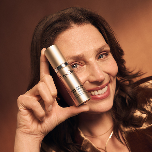 Een glimlachende vrouw die de goudkleurige fles van het Natura Bisse Essential Shock Revolution Collagen Matrix serum naast haar gezicht houdt.