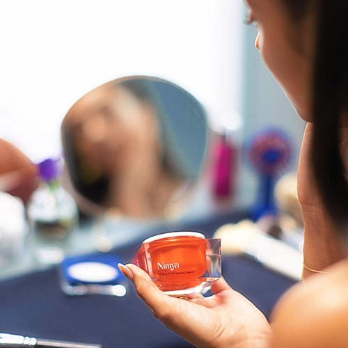 Persoon kijkt in een spiegel en houdt een potje Nimya Where It All Starts Cream vast op een makeuptafel