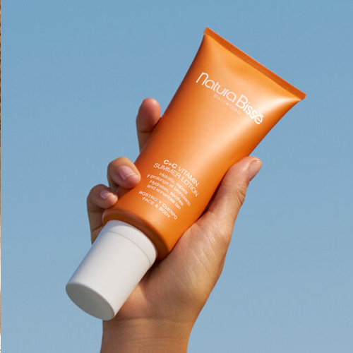 Een hand houdt de oranje tube Natura Bissé C+C Vitamin Summer Lotion omhoog tegen een strakblauwe lucht.Een hand houdt de oranje tube Natura Bissé C+C Vitamin Summer Lotion omhoog tegen een strakblauwe lucht.