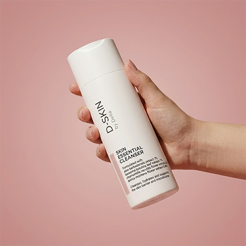 Een hand die een witte fles van de D-SKIN Skin Essential Cleanser schuin vasthoudt tegen een egale roze achtergrond