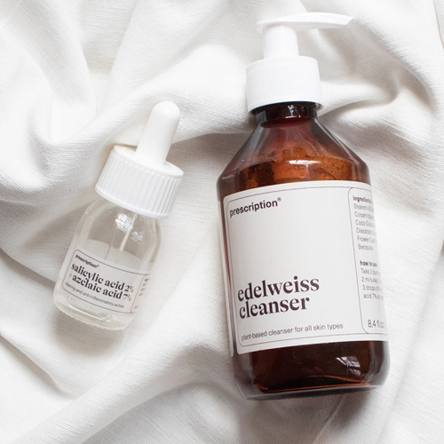 Een bovenaanzicht van een grote amberkleurige fles Prescription edelweiss cleanser naast een kleiner glazen flesje Prescription salicylic acid 2% en azelaic acid 7% serum op een verfrommelde witte stof.