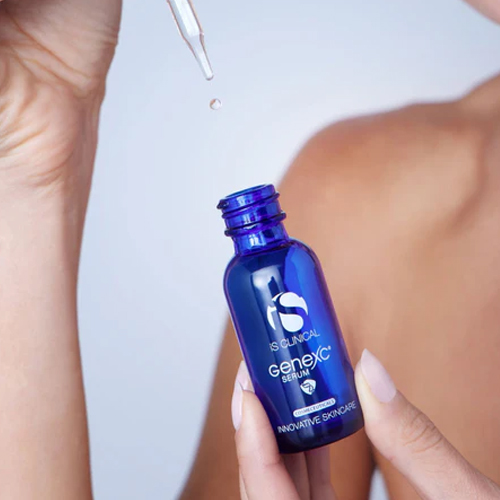 Handen die een donkerblauw glazen flesje van iS Clinical GeneXC Serum vasthouden terwijl er een transparante druppel uit de glazen pipet valt