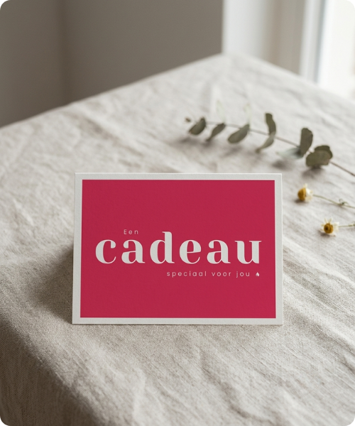 Een staande rode cadeaubon met de witte tekst Een cadeau speciaal voor jou, gepresenteerd op een beige linnen ondergrond. In de achtergrond liggen subtiel enkele gedroogde bloemen.