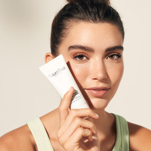 Een glimlachende vrouw houdt de tube van het Natura Bissé Stabilizing Cleansing Mask bij haar gezicht.