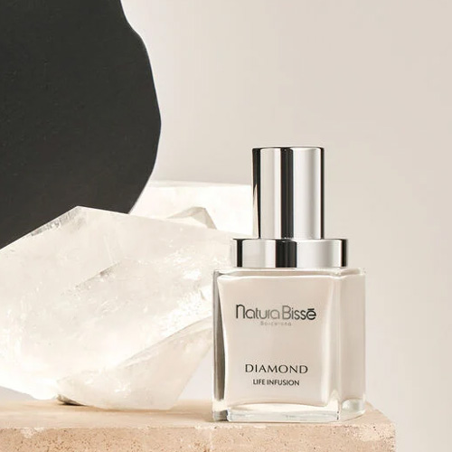 Een flesje Natura Bissé Diamond Life Infusion gezichtsserum gepresenteerd op een stenen plateau naast een groot helder bergkristal tegen een minimalistische achtergrond.