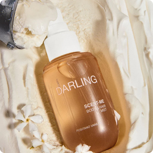 Een glazen flacon van de Darling Scent-Me Body & Hair Mist ligt in een romige bedding van vanille-ijs, met linksboven een ijslepel en onderaan enkele kleine witte bloemetjes