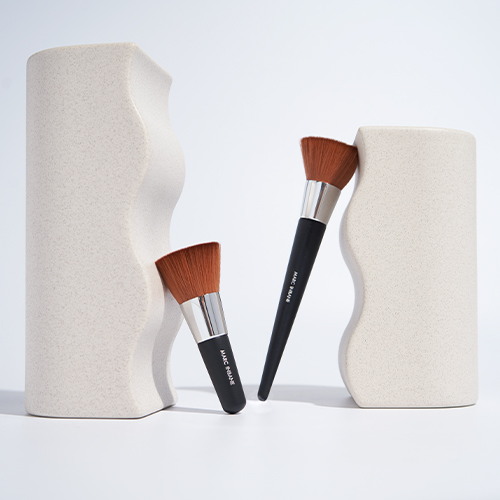 Twee verschillende cosmeticakwasten met bruine haren die nonchalant leunen tegen twee beige golvende design objecten op een witte ondergrond