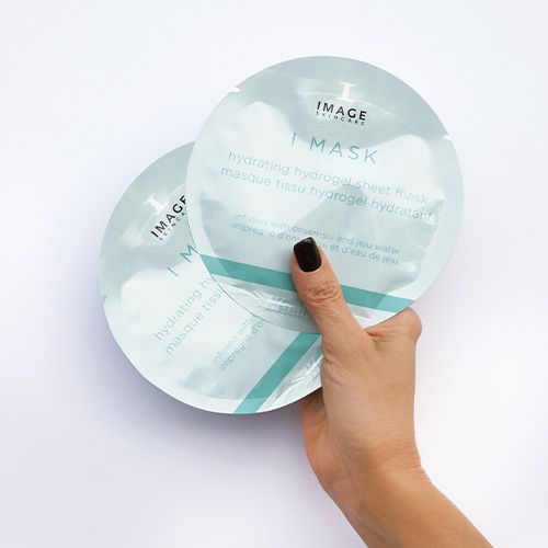 Een hand die twee ronde sachets van het IMAGE Skincare I MASK hydraterende masker vasthoudt tegen een witte achtergrond.