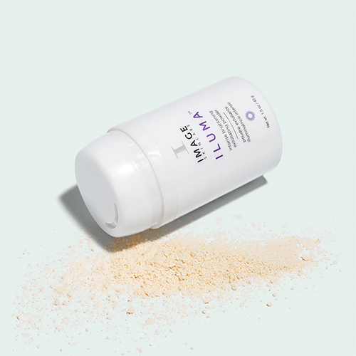 Een witte flacon van Image Skincare Iluma Intense Brightening Exfoliating Powder liggend op een lichtblauwe achtergrond met uitgestrooid poeder.