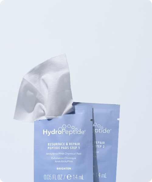 Close-up van twee geopende sachets HydroPeptide Resurface & Repair Peptide Pads waaruit een zachte peeling pad tevoorschijn komt.