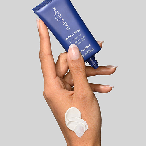 Een hand die een blauwe tube van de HydroPeptide Miracle Mask vasthoudt, met een dot witte maskertextuur op de huid.