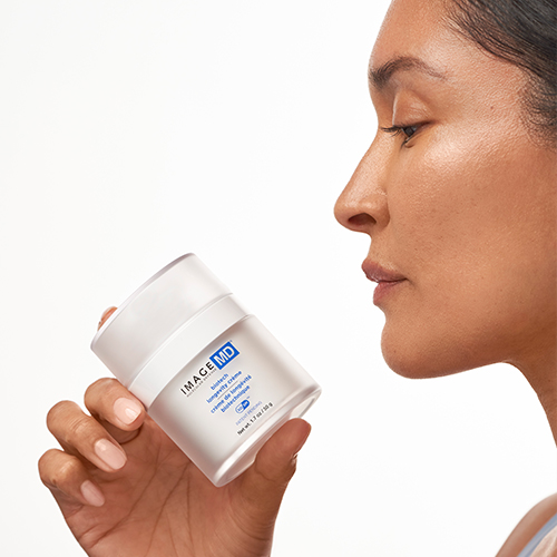 Een vrouw van opzij gefotografeerd die de IMAGE Skincare MD Biotech Longevity Crème pot dicht bij haar gezicht houdt tegen een witte achtergrond.