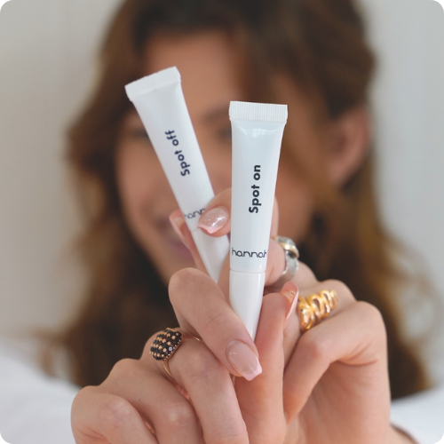 Een vrouw houdt twee witte hannah-tubes vast, de Spot On en Spot Off, met haar handen versierd met goudkleurige ringen.