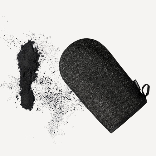 Een bovenaanzicht van de Marc Inbane Exfoliating Mitt scrubhandschoen liggend naast een hoopje fijn zwart houtskoolpoeder op een lichte achtergrond
