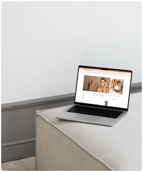 Een geopende zilverkleurige laptop met de homepage van Het Cosmeticahuis op het scherm staat op een grijze gecapitonneerde bank in een kamer met een lichtgrijze muur