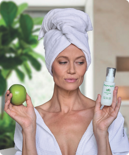 Vrouw met een handdoek op haar hoofd houdt een groene appel in de ene hand en een flesje hannah Cleansing Oil in de andere.