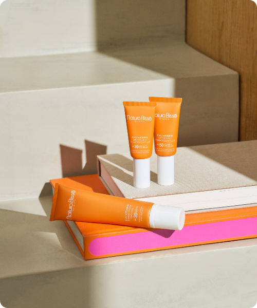 rie oranje tubes van de Natura Bissé C+C Vitamin Dry Touch Sunscreen (SPF 30 en SPF 50) sfeervol opgesteld op en naast boeken op een zonovergoten trap. De afbeelding heeft afgeronde hoeken.