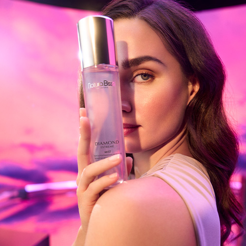 Een vrouw houdt de Diamond Extreme Mist flacon voor haar gezicht tegen een dromerige paars-roze achtergrond.