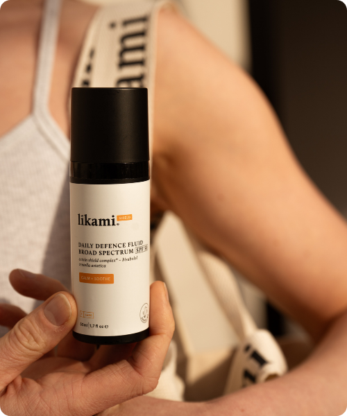 Een persoon houdt een flesje Likami Daily Defence Fluid 'Calm + Soothe' vast tegen een lichte achtergrond.