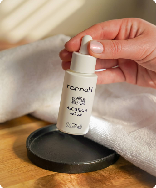 Een lachende jonge vrouw met lang bruin haar in een witte badjas houdt een pipetje boven een flesje hannah Asolution Serum.