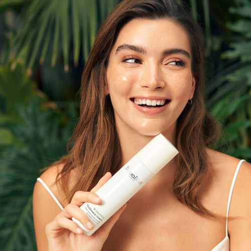 Een lachende vrouw met een gezonde glans op haar gezicht houdt de witte flacon van de Natura Bissé Tolerance Toner vast tegen een achtergrond van groene bladeren.