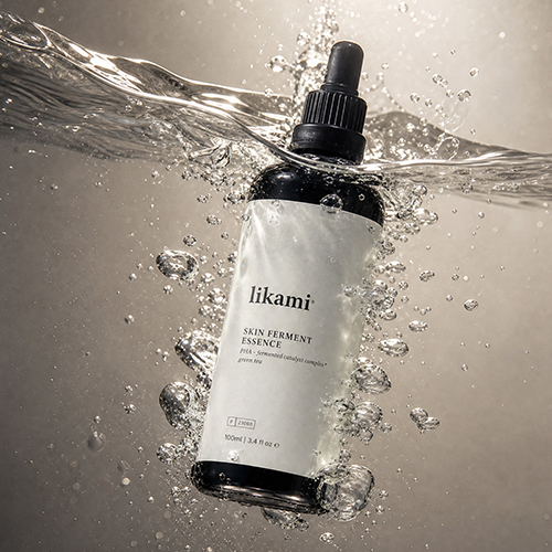 Een dramatische foto van de donkere Likami Skin Ferment Essence fles, ondergedompeld in water tegen een donkerbeige achtergrond. Er zijn tal van luchtbellen rondom de fles en een sterke waterstroming is zichtbaar aan de bovenkant.