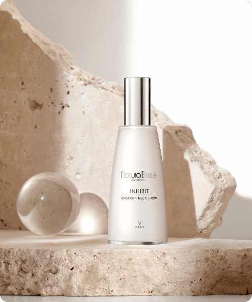 Een flacon Natura Bissé Inhibit Tensolift Neck Serum staand op een stenen voetstuk in een sfeervolle setting met glazen accenten.