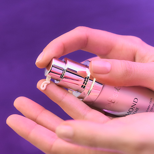 Close-up van handen die de zilveren Diamond Extreme Eye flacon indrukken, waarbij een kleine hoeveelheid oogcrème op de vingertop verschijnt.