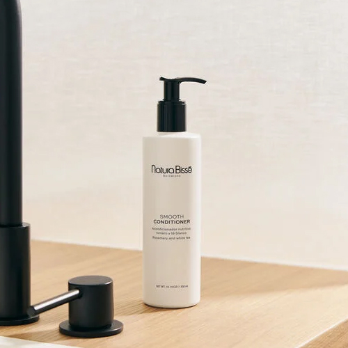 De witte fles Natura Bissé Smooth Conditioner staat op een houten wastafelmeubel naast een moderne zwarte kraan.