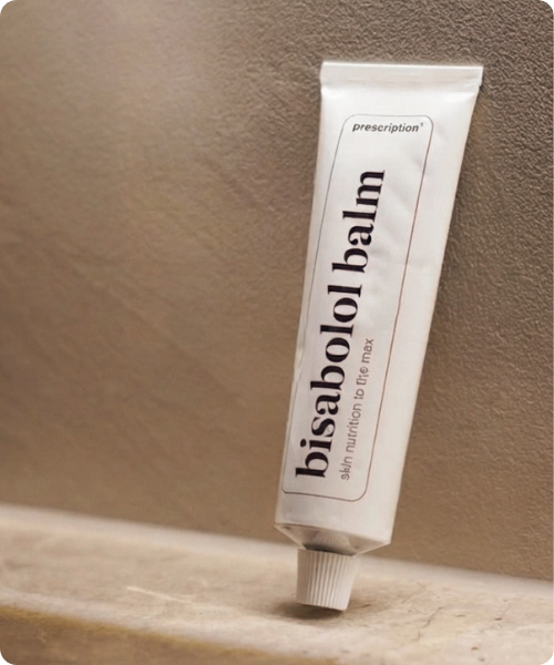 Een witte tube Prescription bisabolol balm staat rechtop tegen een warme beige wand