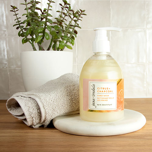 Alt-tekst:  Jane Iredale Citrus + Charcoal Hand Wash gepresenteerd op een houten ondergrond op een marmeren plateau. Naast de vloeibare handzeep liggen een opgerolde handdoek en een groene plant in een witte pot