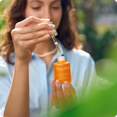 Vrouw in een blauwe blouse houdt een oranje flacon van Natura Bissé C+C Vitamin vast en haalt het doseerpipetje eruit in een natuurlijke, groene buitenomgeving.