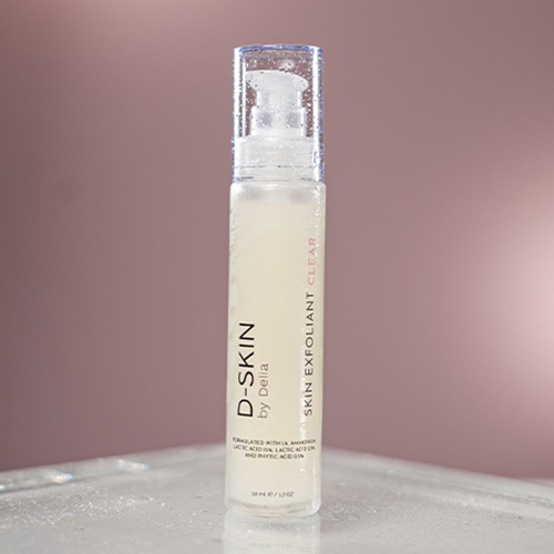 Een flacon D-SKIN Skin Exfoliant Clear bedekt met waterdruppels, staand op een verlicht oppervlak tegen een zachte, roze achtergrond.